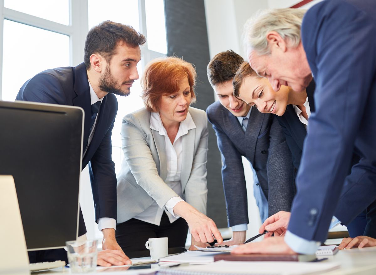 Eine Gruppe von zwei Frauen und drei Männern unterschiedlichen Alters in Business-Kleidung beugt sich über einen Besprechungstisch.